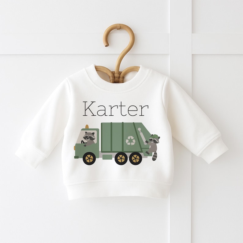 May include: A white baby sweatshirt featuring a green garbage truck design with raccoons and a recycling symbol. The name "Karter" is printed above the truck. The sweatshirt is displayed on a wooden hanger.