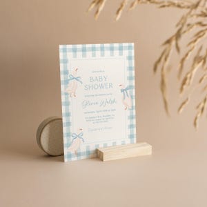 May include: A blue and white gingham baby shower invitation with a blue ribbon and a white goose illustration. The invitation reads "Join us for a Baby Shower Honoring the mommy-to-be Olivia Walsh Saturday, April 15th at 2pm 124 Juniper Way, Boulder, CO RSVP to Lauren by April 1st at [phone number]  Registry at [website]"