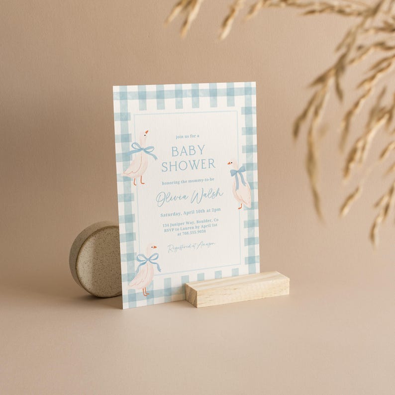 May include: A blue and white gingham baby shower invitation with a blue ribbon and a white goose illustration. The invitation reads "Join us for a Baby Shower Honoring the mommy-to-be Olivia Walsh Saturday, April 15th at 2pm 124 Juniper Way, Boulder, CO RSVP to Lauren by April 1st at [phone number]  Registry at [website]"