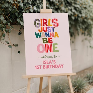 May include: A rectangular sign on a wooden easel with a light pink background. The text reads: GIRLS JUST WANNA BE ONE, - welcome to -, ISLA'S 1ST BIRTHDAY. The words are printed in various colors including purple, yellow, green, red, and pink.