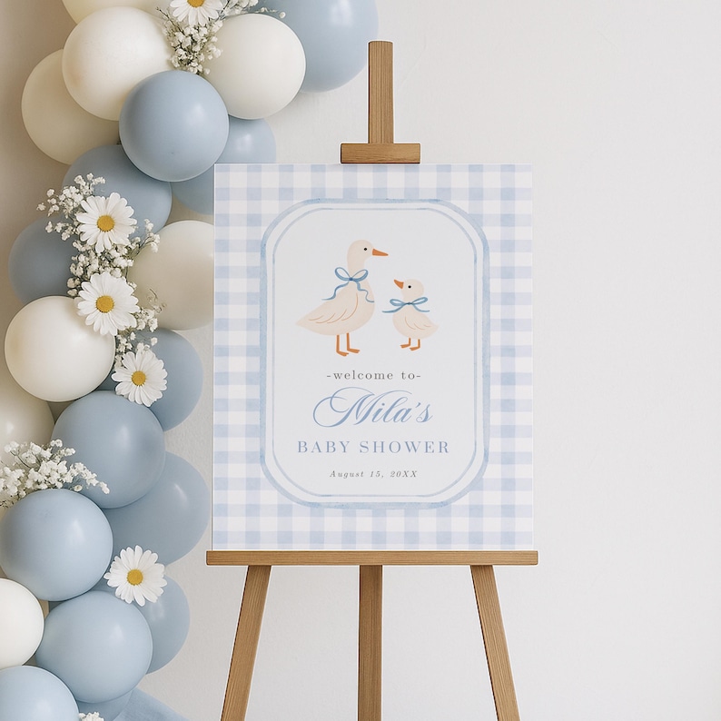 May include: A baby shower sign on a wooden easel. The sign has a blue and white gingham background with a framed illustration of two ducks. The text reads "-welcome to- Nila's BABY SHOWER August 13, 20XX". Balloons and flowers are in the background.
