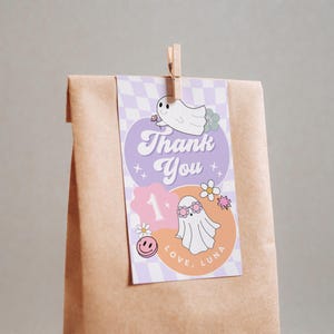May include: A brown paper bag with a decorative tag. The tag features a purple and orange design with the words "Thank You" and "Love, Luna". It includes illustrations of ghosts, flowers, and a smiley face.