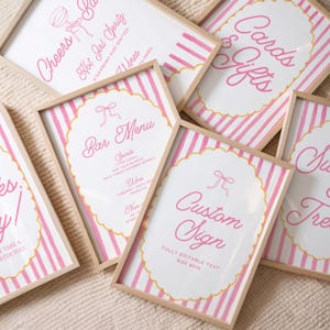 Puede incluir: Una colección de letreros enmarcados con fondos a rayas rosas y blancas y bordes festoneados. Los letreros tienen texto en rosa, incluyendo "Cards & Gifts", "Bar Menu" y "Custom Sign". Los marcos son de madera clara.