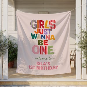 May include: A light pink fabric banner featuring the colorful text "GIRLS JUST WANNA BE ONE" in various patterns and hues. Below, the text reads "welcome to" followed by "ISLA'S 1ST BIRTHDAY" in pink lettering, centered on the hanging backdrop.