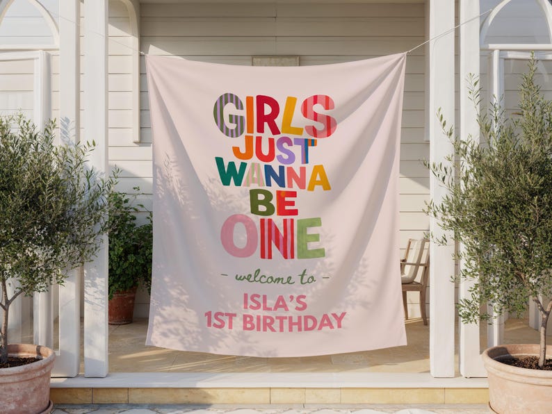 May include: A light pink fabric banner featuring the colorful text "GIRLS JUST WANNA BE ONE" in various patterns and hues. Below, the text reads "welcome to" followed by "ISLA'S 1ST BIRTHDAY" in pink lettering, centered on the hanging backdrop.