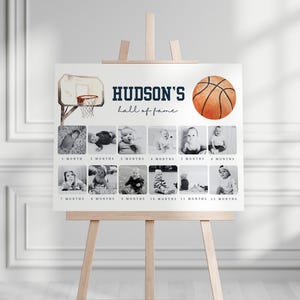 May include: A white poster with a basketball theme for a baby's first year. The poster has a basketball hoop and a basketball with the text "HUDSON'S hall of fame". There are 12 photo spaces for each month of the year.