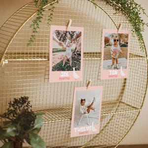 K&ouml;nnte beinhalten: Ein goldfarbenes Drahtregal mit drei rosa Foto-Karten. Jede Karte zeigt ein Baby in verschiedenen Altersstufen mit dem Text "Sechs Monate", "Zehn Monate" und "Zw&ouml;lf Monate".