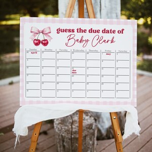 May include: A pink and white baby shower game board on an easel. The sign reads "guess the due date of Baby Clark" with a calendar grid. The top features a bow and cherries. The due date is marked for May 1st.