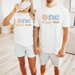 May include: Two people wearing matching white t-shirts with the text "one happy mom" and "one happy dad" in blue and orange. The shirts feature a sun graphic. They are wearing gray shorts.