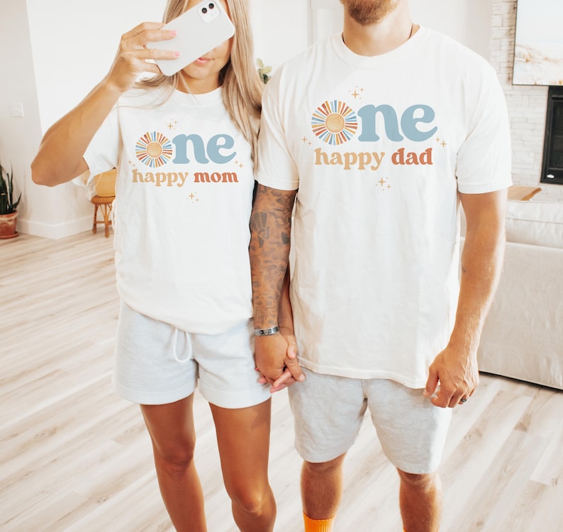 May include: Two people wearing matching white t-shirts with the text "one happy mom" and "one happy dad" in blue and orange. The shirts feature a sun graphic. They are wearing gray shorts.