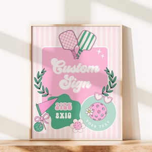 May include: A framed custom sign with a pink and white striped background. The sign features a pink rectangle with the words "Custom Sign" in white script. It includes pickleball paddles, a pickleball, and the text "SIZE 8X10".
