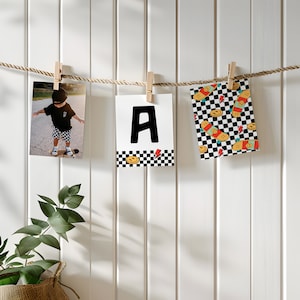 May include: Three white photo cards with black borders are hanging on a string with wooden clothespins. The cards feature a black and white checkered pattern with a smiley face and skateboard graphic. The center card has a large black letter "A" on a white background. The card on the left shows a photo of a young child wearing a black shirt and shorts, standing on a skateboard.