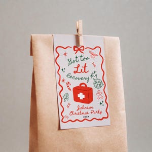 Puede incluir: Una tarjeta blanca con una ilustraci&oacute;n roja y verde de un botiqu&iacute;n de primeros auxilios con el texto "Got too Lit Recovery Kit" y "Johnson Christmas Party 20XX" adjunta a una bolsa de papel marr&oacute;n con una pinza de madera.