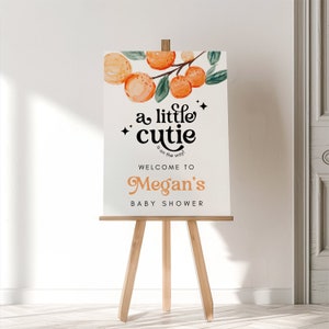 May include: A white sign with orange and green watercolor illustrations of oranges and leaves. The text on the sign reads "a little cutie is on the way! Welcome to Megan's Baby Shower".