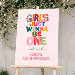 May include: A pink sign on a wooden easel with the text "GIRLS JUST WANNA BE ONE - welcome to - ISLA'S 1ST BIRTHDAY" in colourful letters. The sign is set outdoors with greenery in the background.