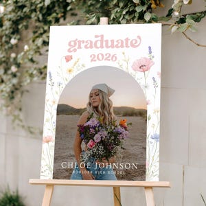 Puede incluir: Un letrero de graduaci&oacute;n sobre un caballete de madera. El letrero presenta el texto "graduate 2026" en escritura rosa, con una foto de una persona sosteniendo flores. El letrero tambi&eacute;n incluye el nombre "Chloe Johnson" y "Boulder High School".