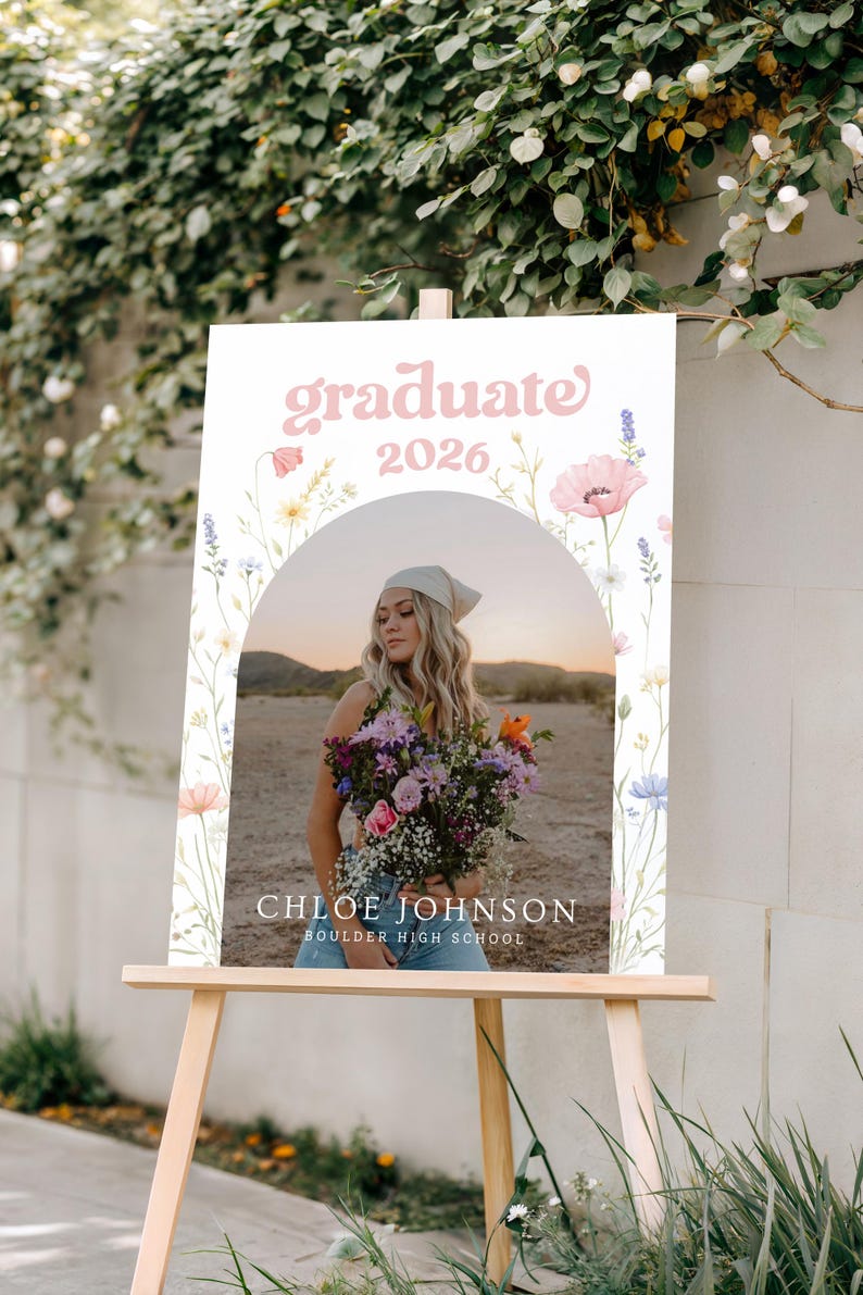 Puede incluir: Un letrero de graduaci&oacute;n sobre un caballete de madera. El letrero presenta el texto "graduate 2026" en escritura rosa, con una foto de una persona sosteniendo flores. El letrero tambi&eacute;n incluye el nombre "Chloe Johnson" y "Boulder High School".