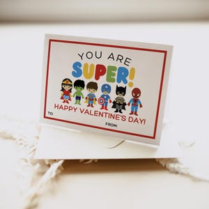 Może przedstawiać: Kartka walentynkowa z czerwoną ramką i tekstem "You are SUPER! Happy Valentine's Day!" Grupa kresk&oacute;wkowych superbohater&oacute;w ustawionych jest w rzędzie u dołu kartki.