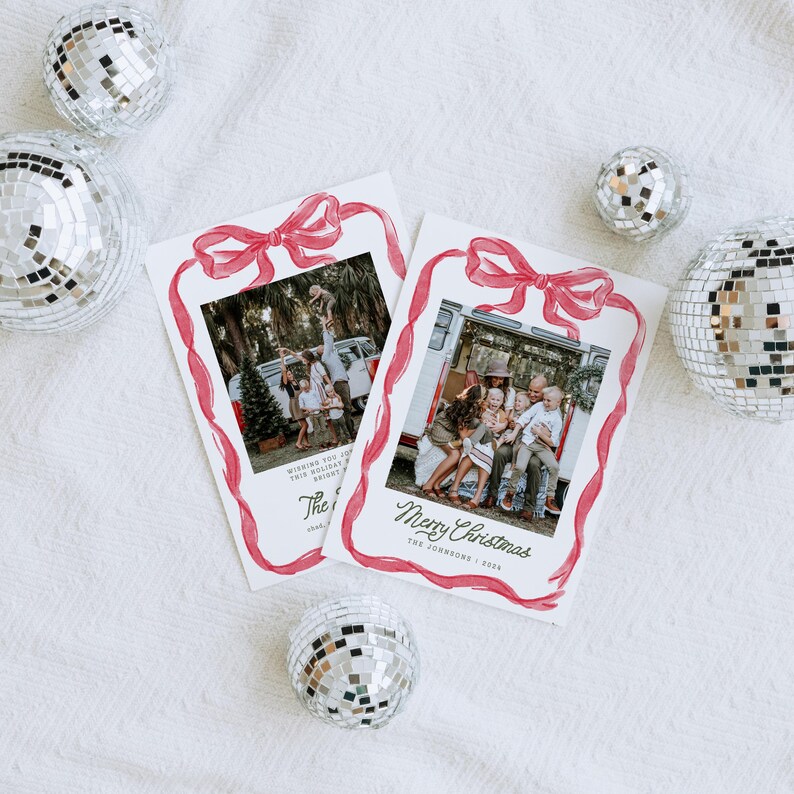 May include: Two Christmas cards featuring family photographs, framed with a red watercolour bow and ribbon design. Silver disco balls are scattered around the cards on a white textured surface. One card says "Merry Christmas".