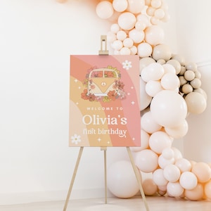 Puede incluir: Un cartel de bienvenida para una fiesta de primer cumplea&ntilde;os con una ilustraci&oacute;n retro de una furgoneta cubierta de flores. El cartel est&aacute; en un caballete de madera y tiene un fondo degradado rosa, naranja y amarillo con estrellas y flores blancas. El texto en el cartel dice "Welcome to Olivia's first birthday".