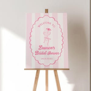 Op de afbeelding: Een roze en witte gestreepte welkomstbord op een houten ezel. Het bord heeft een roze geschulpte ovaal met de woorden "Welcome to" bovenaan, een cocktailillustratie en "Lauren's Bridal Shower" eronder. De datum 09.15.20XX staat onderaan.