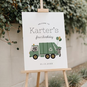 May include: A white sign on a wooden easel with the text "Welcome to Karter's first birthday." A green garbage truck illustration with raccoons and balloons is featured. The date 01.13.20XX is printed below the truck.