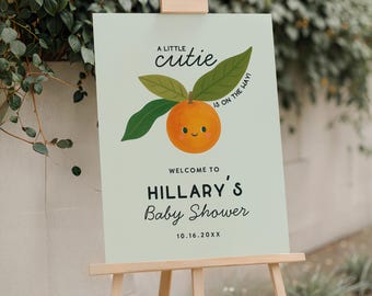 May include: A light green baby shower sign on a wooden easel. The sign reads "A little cutie is on the way!" with an orange illustration. Below, it says "Welcome to Hillary's Baby Shower" and the date.