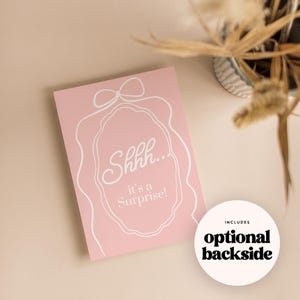 May include: A pink card with white text that reads "Shhh... it's a Surprise!" is displayed. A white decorative outline surrounds the text. A white circle with the words "optional backside" is also visible.