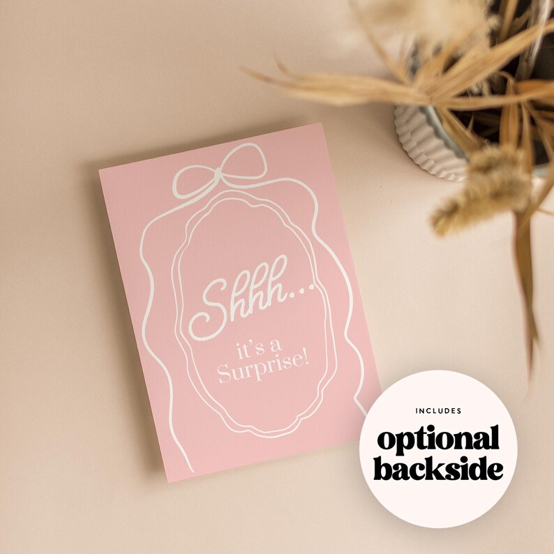 May include: A pink card with white text that reads "Shhh... it's a Surprise!" is displayed. A white decorative outline surrounds the text. A white circle with the words "optional backside" is also visible.