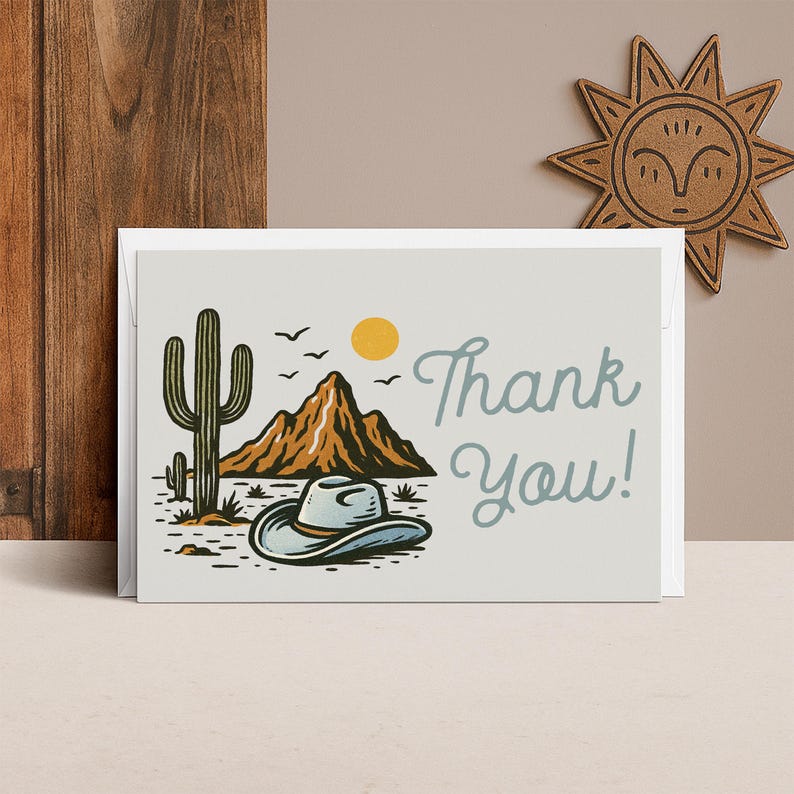 K&ouml;nnte beinhalten: Eine wei&szlig;e Gru&szlig;karte mit einer Illustration zum Thema W&uuml;ste. Das Design umfasst einen Kaktus, Berge, einen Cowboyhut, eine gelbe Sonne und die Worte "Thank You!" in einer Schreibschrift. Eine Holzdekoration in Form einer Sonne befindet sich im Hintergrund.