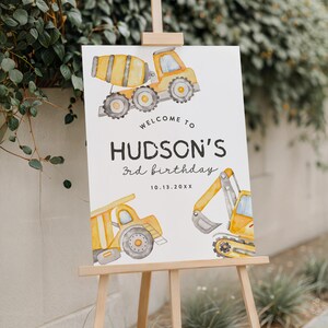 May include: A wooden easel displays a white sign with watercolor illustrations of construction vehicles. The sign reads "Welcome to Hudson's 3rd birthday" with the date 10.13.20XX. The vehicles are yellow and gray.