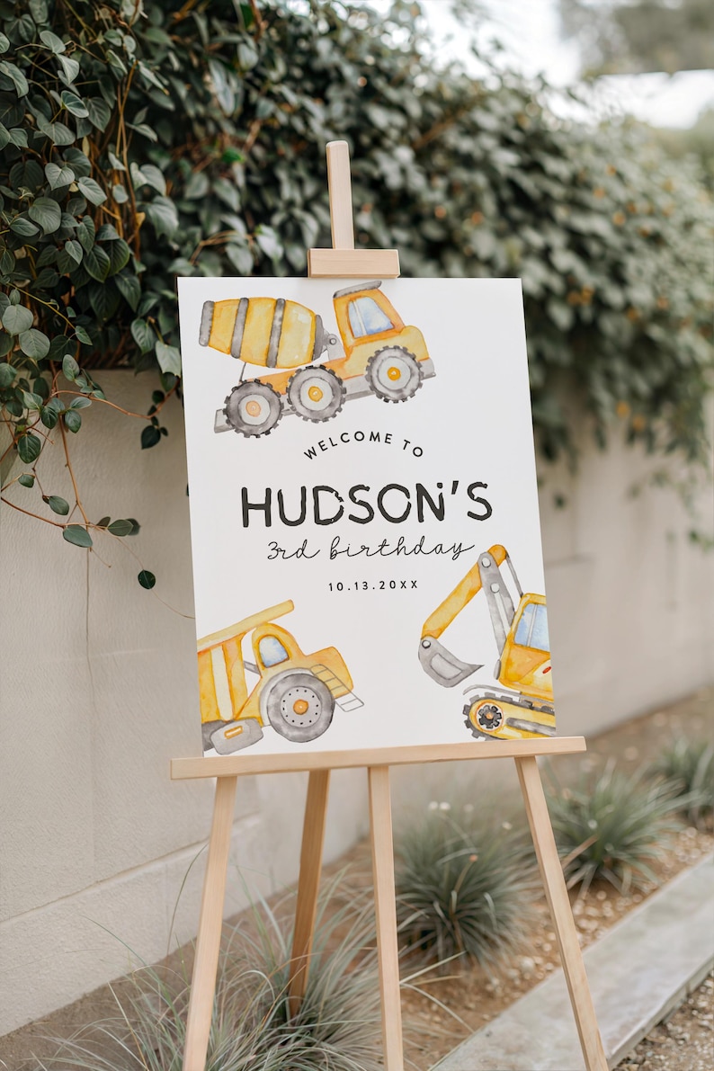 May include: A wooden easel displays a white sign with watercolor illustrations of construction vehicles. The sign reads "Welcome to Hudson's 3rd birthday" with the date 10.13.20XX. The vehicles are yellow and gray.