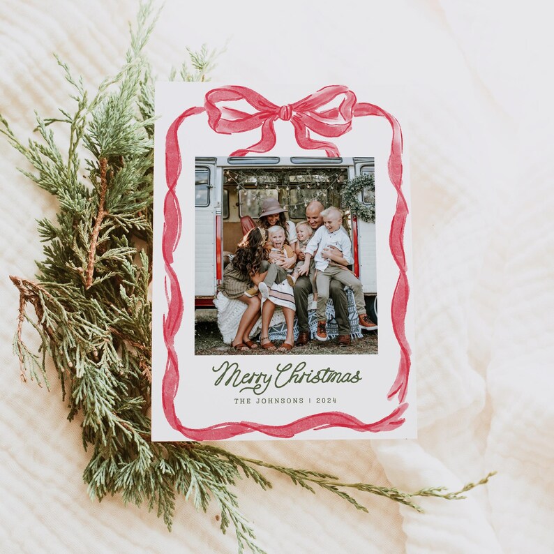May include: A Christmas card featuring a family photograph within a red watercolour ribbon frame. The card says "Merry Christmas" in cursive, with the family name and year below. Green evergreen branches are in the background.
