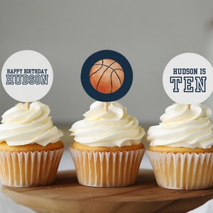 May include: Three cupcakes decorated with basketball-themed toppers. The toppers feature a basketball, the words "Happy Birthday Hudson" and "Hudson is Ten".