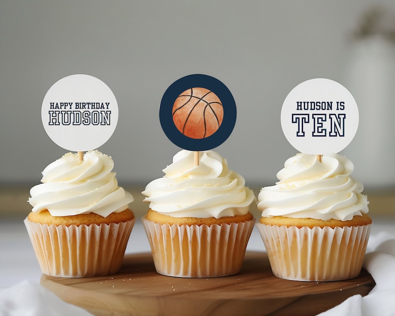 May include: Three cupcakes decorated with basketball-themed toppers. The toppers feature a basketball, the words "Happy Birthday Hudson" and "Hudson is Ten".