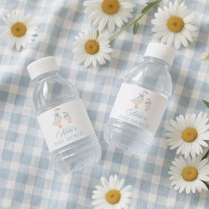 May include: Two clear plastic water bottles with white caps and labels that read "Nila's BABY SHOWER." The labels feature illustrations of ducks with blue bows. The bottles are on a blue and white checkered surface, surrounded by daisies.
