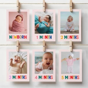 May include: A series of six pink polaroid-style photos clipped to a string with wooden clothespins. Each photo features a baby at different stages, labeled "NEWBORN" to "5 MONTHS" in colorful text. The photos are arranged in two rows.