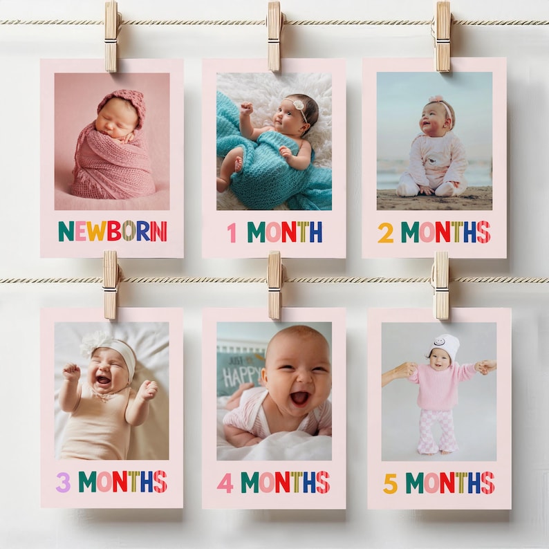 May include: A series of six pink polaroid-style photos clipped to a string with wooden clothespins. Each photo features a baby at different stages, labeled "NEWBORN" to "5 MONTHS" in colorful text. The photos are arranged in two rows.