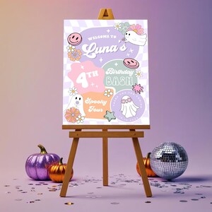 May include: A wooden easel displays a colorful birthday party sign with the text "Welcome to Luna's 4th Birthday Bash". The sign features ghost illustrations, flowers, and smiley faces. A purple glitter pumpkin, an orange pumpkin, and a disco ball are in front of the easel.