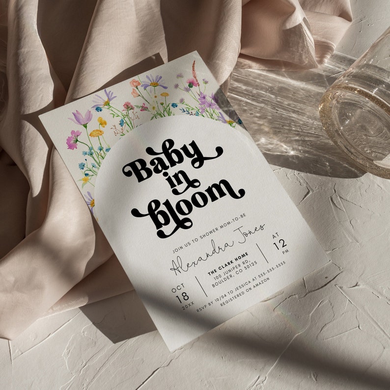 May include: A white baby shower invitation with a watercolor floral design and the text "Baby in Bloom" in a retro font. The invitation includes the date, time, and location of the shower, as well as the name of the mom-to-be, Alexandra Jones.