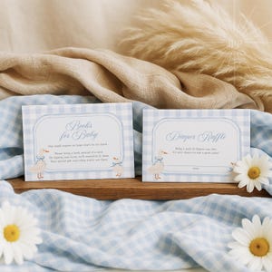May include: Two baby shower cards with blue gingham borders. One card reads "Books for Baby" and the other "Diaper Raffle". Both cards feature a cartoon duck illustration. The cards are on a wooden tray with daisies.