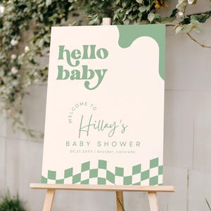 May include: A rectangular welcome sign for a baby shower featuring a cream background with sage green accents. The text reads hello baby, welcome to Hillay's baby shower, 05.27.20XX, and boulder, colorado. The bottom edge has a sage green checkered pattern.
