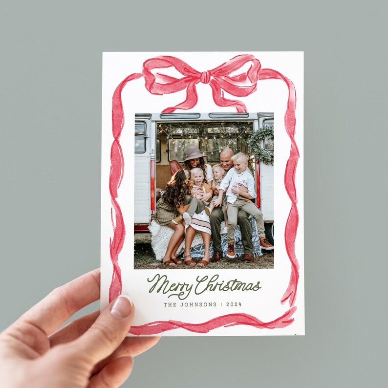 May include: A Christmas card with a family photo framed by a red watercolour bow and ribbon design. The card reads "Merry Christmas" in cursive, with "The Johnsons | 2024" underneath. The card is held in a hand.