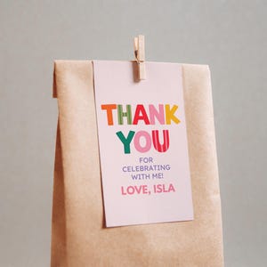 May include: A light pink rectangular paper tag attached to a brown paper bag with a wooden clothespin. The tag features the colorful text THANK YOU, FOR CELEBRATING WITH ME!, and LOVE, ISLA in various fonts and bright colors including orange, red, and green.