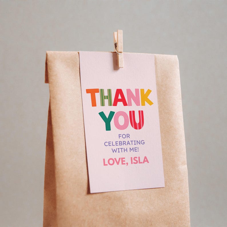 May include: A light pink rectangular paper tag attached to a brown paper bag with a wooden clothespin. The tag features the colorful text THANK YOU, FOR CELEBRATING WITH ME!, and LOVE, ISLA in various fonts and bright colors including orange, red, and green.