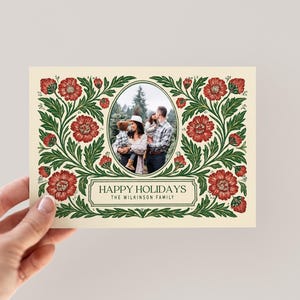 May include: A festive holiday card with a family photograph at its centre. The card has a cream-coloured background and is framed by a floral border of red flowers and green foliage. The text reads "HAPPY HOLIDAYS" and the family name.