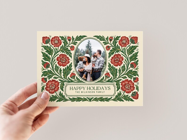 May include: A festive holiday card with a family photograph at its centre. The card has a cream-coloured background and is framed by a floral border of red flowers and green foliage. The text reads "HAPPY HOLIDAYS" and the family name.