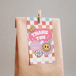 May include: A pink and purple thank you tag with a checkered background. The tag has a pink banner with the text "THANK YOU" in white. There are smiley faces and flowers on the tag. The tag is attached to a brown paper bag with a wooden clothespin.