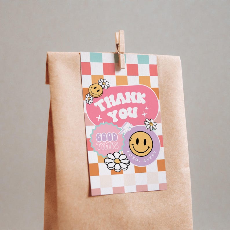 May include: A pink and purple thank you tag with a checkered background. The tag has a pink banner with the text "THANK YOU" in white. There are smiley faces and flowers on the tag. The tag is attached to a brown paper bag with a wooden clothespin.