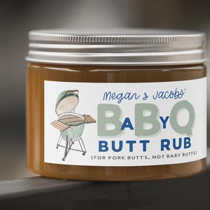 May include: A jar of BBQ butt rub with a silver lid and a white label. The label features the text "Megan & Jacobs BABYQ BUTT RUB" and an illustration of a grill. The jar is filled with a brown substance.